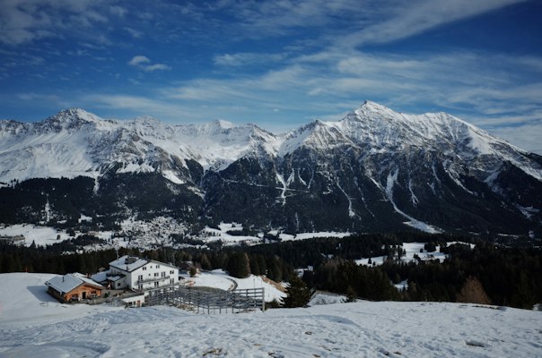 A timber hut and hotel buildings on a snowy terrace facing the snow-covered Lenzerheide-side range