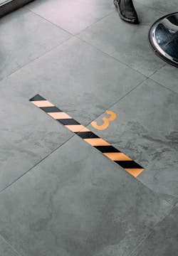 A gray tiled floor with a yellow number three and a black and yellow striped line. Part of a chair or stool leg and a shoe are visible at the edges of the image.