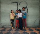Four children stand closely together against a metal, light green shutter with graffiti. The floor features a black and orange checkered pattern. The children are dressed casually, with colorful outfits that include sweaters, pants, and slippers.