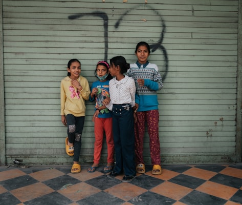 Four children stand closely together against a metal, light green shutter with graffiti. The floor features a black and orange checkered pattern. The children are dressed casually, with colorful outfits that include sweaters, pants, and slippers.