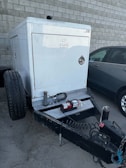 A trailer-mounted generator is positioned in a parking area with a grey car nearby. The generator has a metallic enclosure with visible branding. Attached accessories include coiled cables and a fire extinguisher placed on the platform. The background features a tiled concrete wall.