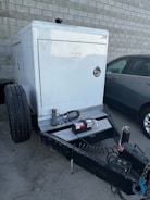 A trailer-mounted generator is positioned in a parking area with a grey car nearby. The generator has a metallic enclosure with visible branding. Attached accessories include coiled cables and a fire extinguisher placed on the platform. The background features a tiled concrete wall.