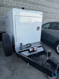 A trailer-mounted generator is positioned in a parking area with a grey car nearby. The generator has a metallic enclosure with visible branding. Attached accessories include coiled cables and a fire extinguisher placed on the platform. The background features a tiled concrete wall.