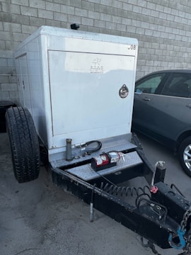 A trailer-mounted generator is positioned in a parking area with a grey car nearby. The generator has a metallic enclosure with visible branding. Attached accessories include coiled cables and a fire extinguisher placed on the platform. The background features a tiled concrete wall.