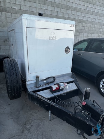 A trailer-mounted generator is positioned in a parking area with a grey car nearby. The generator has a metallic enclosure with visible branding. Attached accessories include coiled cables and a fire extinguisher placed on the platform. The background features a tiled concrete wall.