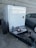 A trailer-mounted generator is positioned in a parking area with a grey car nearby. The generator has a metallic enclosure with visible branding. Attached accessories include coiled cables and a fire extinguisher placed on the platform. The background features a tiled concrete wall.