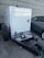 A trailer-mounted generator is positioned in a parking area with a grey car nearby. The generator has a metallic enclosure with visible branding. Attached accessories include coiled cables and a fire extinguisher placed on the platform. The background features a tiled concrete wall.