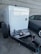 A trailer-mounted generator is positioned in a parking area with a grey car nearby. The generator has a metallic enclosure with visible branding. Attached accessories include coiled cables and a fire extinguisher placed on the platform. The background features a tiled concrete wall.