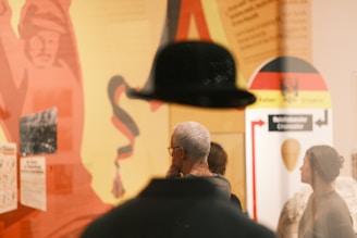 A scene in what appears to be a museum or exhibition setting, with people standing in front of colorful displays. The background features various graphics and texts with predominantly red, yellow, and black colors. Reflections and shadows add an abstract element to the composition.