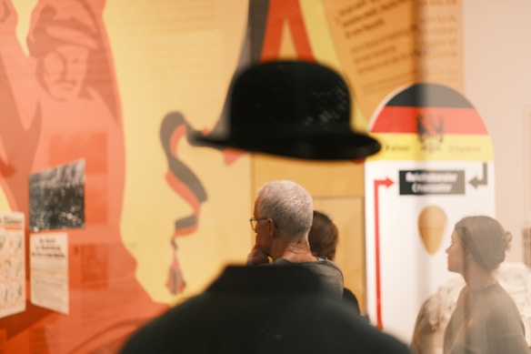 A scene in what appears to be a museum or exhibition setting, with people standing in front of colorful displays. The background features various graphics and texts with predominantly red, yellow, and black colors. Reflections and shadows add an abstract element to the composition.