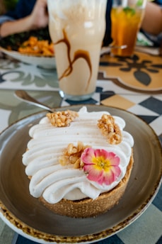 A beautifully presented dessert with elaborately piped whipped cream, garnished with nuts and a small pink flower on top of a crumbly crust. In the background, there are drinks, including an iced coffee with cream, beside plates with other food items. The table has a colorful, patterned design.