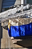 A balcony with railings is adorned with white fabric strips blowing in the wind. Below the strips are two large pieces of fabric, one yellow and one blue, draped over the railing. The background features a textured stone wall and a partially visible window with closed shutters.