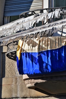 A balcony with railings is adorned with white fabric strips blowing in the wind. Below the strips are two large pieces of fabric, one yellow and one blue, draped over the railing. The background features a textured stone wall and a partially visible window with closed shutters.
