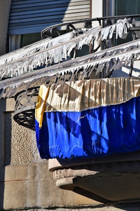 A balcony with railings is adorned with white fabric strips blowing in the wind. Below the strips are two large pieces of fabric, one yellow and one blue, draped over the railing. The background features a textured stone wall and a partially visible window with closed shutters.
