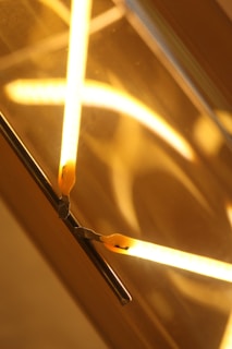 Bright yellow neon lights are arranged in an abstract pattern against a reflective surface, creating an intersecting design. The warm glow contrasts against the darker background, highlighting the glass and metal elements.