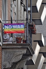 A rainbow-colored flag with the words 'NO WAR' hangs from the railing of a balcony. The building features brick and stone elements, with potted plants visible on the balcony. The atmosphere conveys a message of peace and anti-war sentiment.