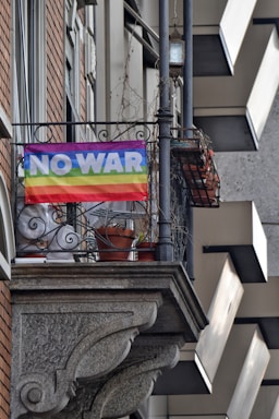 A rainbow-colored flag with the words 'NO WAR' hangs from the railing of a balcony. The building features brick and stone elements, with potted plants visible on the balcony. The atmosphere conveys a message of peace and anti-war sentiment.
