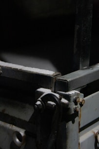 A close-up view of a metal structure with bolts and a handle. The surface appears to have a layer of dust or corrosion. The lighting is dim, creating strong shadows and highlighting the texture of the metal.