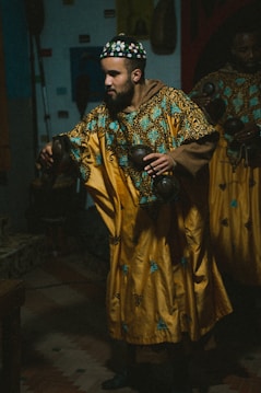 A person wearing a traditional, ornate garment with intricate turquoise and black embroidery. They hold musical instruments in both hands, with a focused expression. The setting appears dimly lit, suggesting an intimate or ceremonial atmosphere.