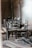 A collection of wooden chairs is arranged in a seemingly haphazard manner in a rustic, dimly lit wooden room. The chairs are a mix of styles and colors, placed amid scattered wooden planks and boards, creating an abandoned or unfinished atmosphere.