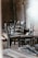 A collection of wooden chairs is arranged in a seemingly haphazard manner in a rustic, dimly lit wooden room. The chairs are a mix of styles and colors, placed amid scattered wooden planks and boards, creating an abandoned or unfinished atmosphere.