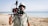 A person stands on a sandy beach near a calm ocean, holding a professional camera setup. They are wearing a floral shirt, a bucket hat, and sunglasses. The background shows a clear sky and a serene seascape.