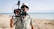 A person stands on a sandy beach near a calm ocean, holding a professional camera setup. They are wearing a floral shirt, a bucket hat, and sunglasses. The background shows a clear sky and a serene seascape.