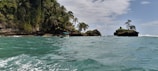 A scenic coastal area with a turquoise sea in the foreground. Lush green vegetation and palm trees cover a rocky island. A small boat with a canopy and a few passengers is navigating the water near the shoreline. The sky is partly cloudy, creating a serene atmosphere.
