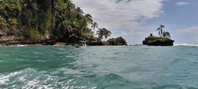 A scenic coastal area with a turquoise sea in the foreground. Lush green vegetation and palm trees cover a rocky island. A small boat with a canopy and a few passengers is navigating the water near the shoreline. The sky is partly cloudy, creating a serene atmosphere.