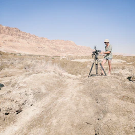 A person in boots walking through rugged desert terrain with a camera in hand.