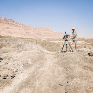 A person in boots walking through rugged desert terrain with a camera in hand.