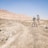 In a rugged, arid landscape with rocky terrain and distant barren hills under a clear blue sky, an individual wearing a hat and shorts operates a professional camera mounted on a tripod.