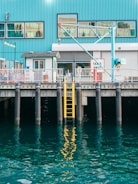A waterfront scene featuring a blue industrial building with large windows and a crane. A yellow ladder is attached to the dock, descending into the reflective water. The dock has signage indicating 'Taxies Only'.