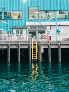 A waterfront scene featuring a blue industrial building with large windows and a crane. A yellow ladder is attached to the dock, descending into the reflective water. The dock has signage indicating 'Taxies Only'.