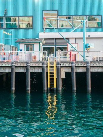 A waterfront scene featuring a blue industrial building with large windows and a crane. A yellow ladder is attached to the dock, descending into the reflective water. The dock has signage indicating 'Taxies Only'.