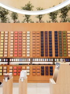 A stylish display wall featuring a wide array of colorful coffee pods arranged in a grid pattern. The pods are of various colors such as red, orange, gold, and navy blue, set against a light wooden backdrop. The upper part showcases artificial trees and a modern, circular light fixture overhead. In the foreground, a sleek checkout counter and stands with products add to the modern retail atmosphere.