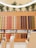 A stylish display wall featuring a wide array of colorful coffee pods arranged in a grid pattern. The pods are of various colors such as red, orange, gold, and navy blue, set against a light wooden backdrop. The upper part showcases artificial trees and a modern, circular light fixture overhead. In the foreground, a sleek checkout counter and stands with products add to the modern retail atmosphere.