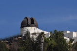 An observatory with a domed telescope structure sits atop a hill surrounded by trees. The clear blue sky provides a contrasting backdrop. The facility is a mix of modern and classical architectural elements, with large windows and white walls.