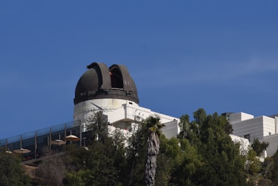 An observatory with a domed telescope structure sits atop a hill surrounded by trees. The clear blue sky provides a contrasting backdrop. The facility is a mix of modern and classical architectural elements, with large windows and white walls.