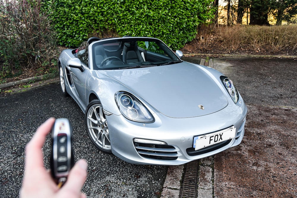 a person holding a cell phone in front of a silver sports car