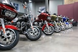A row of motorcycles is neatly lined up indoors on a polished concrete floor. The motorcycles are of various colors, including red, black, gold, and blue, and have large, prominent wheels. The background features a gray wall with framed images and exposed brick, giving the space a modern, industrial feel.
