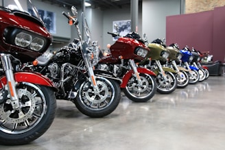 A row of motorcycles is neatly lined up indoors on a polished concrete floor. The motorcycles are of various colors, including red, black, gold, and blue, and have large, prominent wheels. The background features a gray wall with framed images and exposed brick, giving the space a modern, industrial feel.