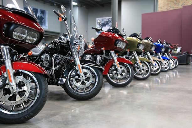 A row of motorcycles is neatly lined up indoors on a polished concrete floor. The motorcycles are of various colors, including red, black, gold, and blue, and have large, prominent wheels. The background features a gray wall with framed images and exposed brick, giving the space a modern, industrial feel.