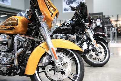 Two Harley-Davidson motorcycles are parked side by side in a showroom. The one in the foreground is golden with flame accents, featuring a large front fairing and shiny chrome parts. The motorcycle behind it is black, similarly styled and polished. The background consists of showroom features like posters and bike-related merchandise.