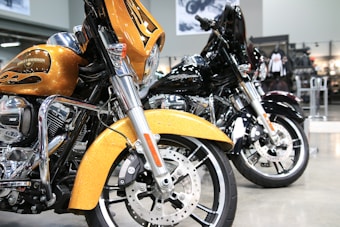 Two Harley-Davidson motorcycles are parked side by side in a showroom. The one in the foreground is golden with flame accents, featuring a large front fairing and shiny chrome parts. The motorcycle behind it is black, similarly styled and polished. The background consists of showroom features like posters and bike-related merchandise.