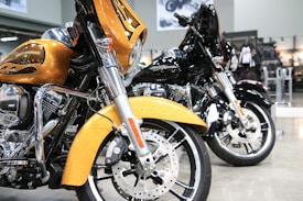 Two Harley-Davidson motorcycles are parked side by side in a showroom. The one in the foreground is golden with flame accents, featuring a large front fairing and shiny chrome parts. The motorcycle behind it is black, similarly styled and polished. The background consists of showroom features like posters and bike-related merchandise.
