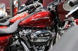 A close-up view of a red motorcycle with chrome details and branding visible on the fuel tank. The bike is positioned indoors, possibly in a showroom. Surrounding motorcycles are partially visible, adding depth to the scene.