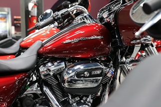 A close-up view of a red motorcycle with chrome details and branding visible on the fuel tank. The bike is positioned indoors, possibly in a showroom. Surrounding motorcycles are partially visible, adding depth to the scene.