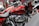 A close-up view of a red motorcycle with chrome details and branding visible on the fuel tank. The bike is positioned indoors, possibly in a showroom. Surrounding motorcycles are partially visible, adding depth to the scene.