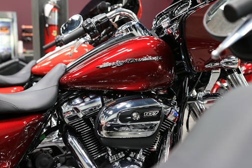 A close-up view of a red motorcycle with chrome details and branding visible on the fuel tank. The bike is positioned indoors, possibly in a showroom. Surrounding motorcycles are partially visible, adding depth to the scene.